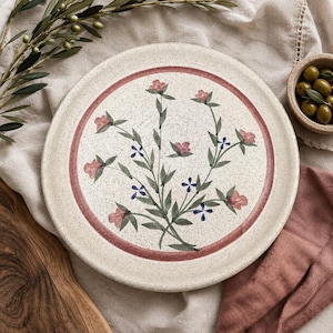 May include: A round ceramic plate with a speckled cream finish, featuring a central hand-painted botanical motif of pink flowers and green leaves enclosed by a thin red circular border. The plate has a smooth, flat surface design.