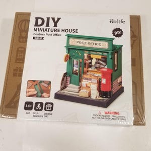 May include: A box for a DIY miniature house kit, featuring a green and brown post office building with a red mailbox. The box has the text "DIY Miniature House Century Post Office D5037" and "Rolife DIY" on it. The box also has a warning label that says "CHOKING HAZARD - SMALL PARTS. NOT FOR CHILDREN UNDER 3 YEARS."