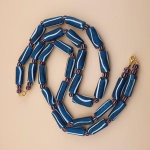 Puede incluir: Un collar con cuentas azul oscuro en forma de barril con rayas blancas. Las cuentas están unidas con cuentas más pequeñas de color púrpura rojizo. Tiene un cierre dorado.