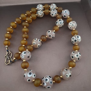 May include: A necklace with brown and speckled pink beads. The necklace has a gold clasp and small gold beads between the larger beads. The speckled beads have a white background with black dots. The necklace is displayed on a brown surface.