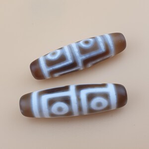 May include: Two elongated, brown and white dzi beads. Each bead features a repeating geometric pattern in white against a brown background. The beads have rounded ends and a smooth texture, suitable for jewelry making or collecting.