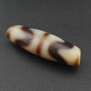 May include: A polished, oblong dzi bead with a cream-colored base and swirling patterns of dark brown. The bead has a smooth, glossy finish and is likely made of agate or a similar stone.
