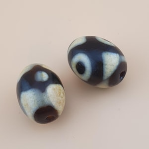 May include: Two oval dzi beads. The beads are mainly black with cream patterns. Each bead has a hole through its centre. The beads are smooth and polished, with a glossy finish. They are set against a light beige background.