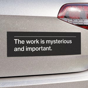 May include: A black rectangular car magnet with the text "The work is mysterious and important" in white. The magnet is affixed to the rear of a silver car, near the taillight.
