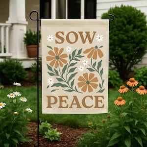 Könnte beinhalten: Eine beige Gartenflagge mit den Worten "SOW PEACE" in Braun, umgeben von braunen und weißen Blumen und grünen Blättern. Die Flagge wird in einem Garten mit grünem Gras und Blumen gezeigt.