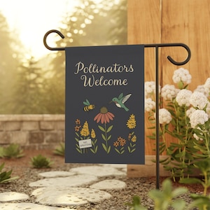 Könnte beinhalten: Eine Gartenflagge mit den Worten "Pollinators Welcome" in einer dekorativen Schriftart. Die Flagge zeigt Illustrationen einer Biene, eines Kolibris und verschiedener Blumen. Ein kleines Schild lautet "No Spraying". Die Flagge wird auf einem schwarzen Metallständer angezeigt.