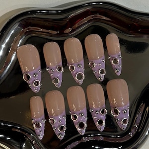 May include: A set of ten artificial nails. Each nail has a nude base with a lavender tip, adorned with silver-colored metal rings and studs. The nails are arranged on a silver-colored tray.