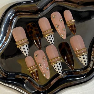Op de afbeelding: Een set van tien kunstnagels in verschillende ontwerpen, waaronder effen roze, luipaardprint en wit met zwarte stippen. Sommige nagels hebben gouden ringen en strass-steentjes. De nagels worden gepresenteerd op een zilveren dienblad.