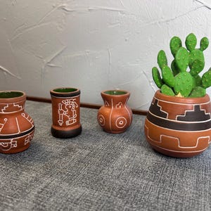 May include: Four terracotta pottery pieces. Three small vases with white geometric designs and a green interior. A larger pot with a cactus plant, featuring a brown and black geometric pattern and white accents. The items are on a gray surface.