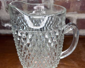 May include: A clear glass pitcher with a diamond pattern. The pitcher has a handle and a wide mouth.