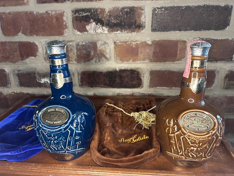 May include: Two ceramic bottles of Royal Salute Scotch whisky. The bottle on the left is blue with gold accents and the bottle on the right is brown with gold accents. Both bottles have a label with the words "Royal Salute" and "Scotch Whisky".