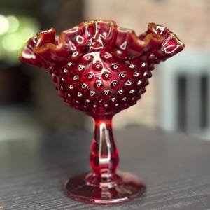 May include: A deep red glass compote dish featuring a hobnail texture across the bowl and a ruffled, crimped rim. The piece stands on a pedestal base with a circular foot, showcasing a translucent finish that catches the light throughout.