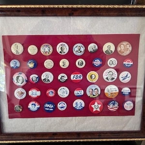 May include: Framed collection of vintage political campaign buttons. The buttons, in various sizes, feature portraits, slogans, and patriotic imagery. The buttons are arranged on a red background within a wooden frame with a gold trim.