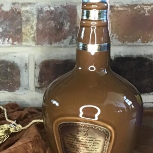 May include: A brown ceramic bottle with a silver top and a label that reads "By Royal Command, Supplying the Royal Navy,  is making annually one gift,  the presentation of the  'Chivas Regal'  to the  'Royal Navy'  in commemoration of her  Majesty the Queen's  Golden Jubilee, 1977."