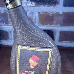 May include: A vintage liquor bottle with a gold-colored cap and a label featuring a painting of a boy with cherries. The bottle has a textured, dark brown finish with gold accents. The label reads "Boy with Cherries" by Edouard Manet.