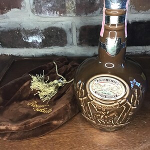May include: A brown ceramic bottle of Royal Salute Blended Scotch Whisky with a gold label and a gold tassel on a brown velvet bag. The bottle is decorated with a gold design of a stag and a lion.