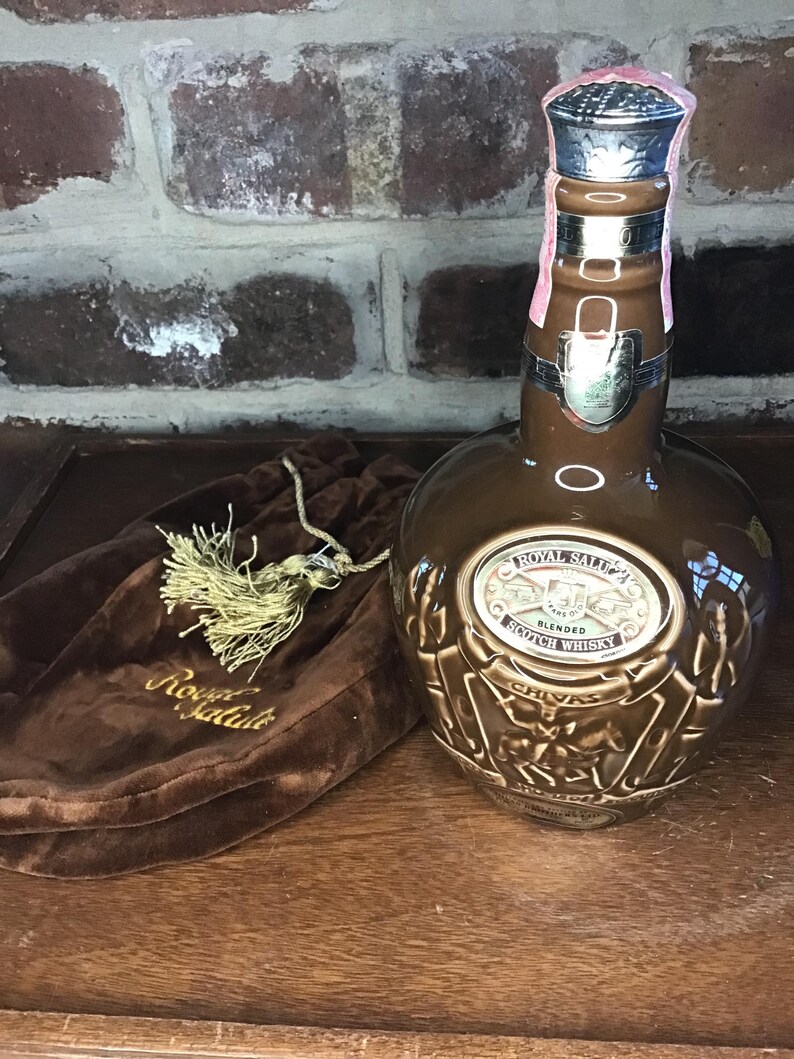 May include: A brown ceramic bottle of Royal Salute Blended Scotch Whisky with a gold label and a gold tassel on a brown velvet bag. The bottle is decorated with a gold design of a stag and a lion.