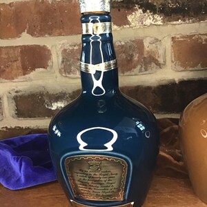 May include: A blue ceramic bottle of Chivas Regal Scotch Whisky with a gold label and a silver cap. The label reads "By an ancient custom, recognizing the Royal Navy, a whisky of beauty and rare quality in the prerogative of the Sovereign. This bottle of Chivas Regal Whisky created in the 1980s for the 125th Anniversary of the Royal Navy, is presented to Her Majesty Queen Elizabeth II."