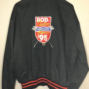 May include: A black bomber jacket with red and black striped ribbed cuffs and hem. The back features an embroidered patch with the text ROD & Custom AMERICRUISE Tour '95 in white, red, yellow, and blue colors, centered on the upper back panel.