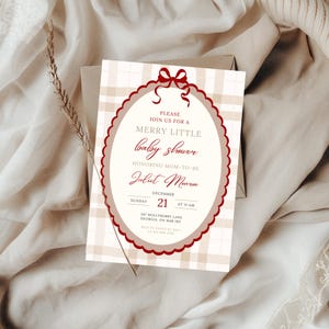 May include: A baby shower invitation with a plaid background and a decorative oval frame. The invitation text is in burgundy and includes the words "Merry Little baby shower" and the name "Juliet Monroe". The date is December 21st.