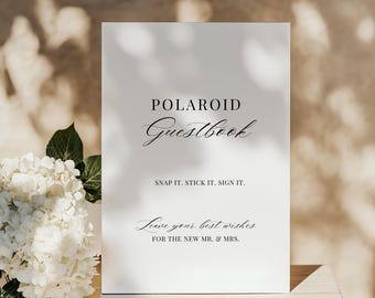 May include: A white rectangular sign with black text that reads: POLAROID Guestbook, SNAP IT. STICK IT. SIGN IT., Leave your best wishes FOR THE NEW MR. & MRS. The sign features a mix of serif and script fonts and is displayed on a light surface.