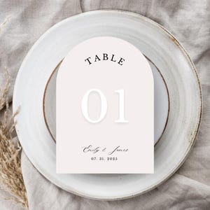 May include: A table number card with an arched top, displaying the word "TABLE" above the number "01". Below, the names "Emily & James" and the date "07. 31. 2025" are printed. The card is set on a white plate with a brown rim.