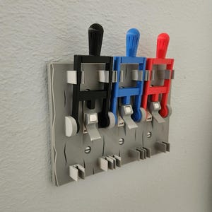 May include: A triple light switch cover plate featuring a gray base with a textured finish. It includes three vertical lever-style toggles in black, blue, and red, each with light gray accents and mounting hardware visible on the faceplate.
