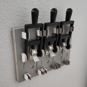Pode incluir: Uma placa de cobertura para interruptor triplo com três extensões de alavanca pretas de estilo industrial fixadas numa base retangular cinzenta. O mecanismo inclui componentes de plástico branco e cinzento sobre os interruptores.
