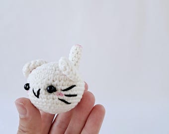 Peut inclure: Tête de chat blanche crochetée avec des yeux noirs, des moustaches et un nez rose. Le chat a une oreille dressée avec une pointe rose et deux petites oreilles. Le chat fait main est un design charmant et fantaisiste.