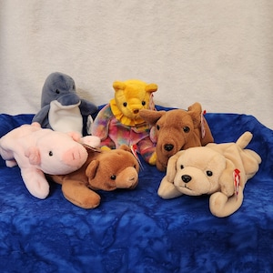 May include: A group of six plush toys, including a pink pig, a brown bear, a yellow bear, a blue dolphin, a brown dog, and a beige dog. The toys are arranged on a blue fabric background.