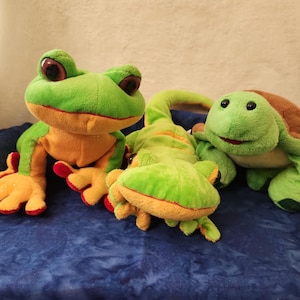 May include: Three plush toys: a green frog with orange accents and red feet, a green lizard with orange stripes, and a green turtle with a brown shell. The toys are on a blue textured surface.