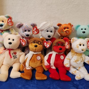 May include: A collection of nine plush teddy bears featuring various colors and accessories. One white bear displays the text I Love New York. Several bears wear hats, scarves, or goggles, and each includes a red heart-shaped Ty tag attached to the ear.