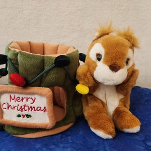May include: A plush Christmas tree trunk with a "Merry Christmas" sign and a brown squirrel toy. The tree trunk is green with colorful lights and a tan interior. The squirrel is brown and white.