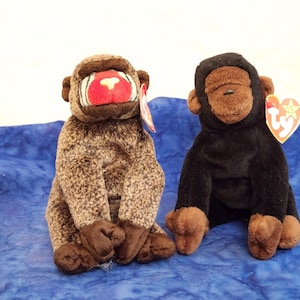 May include: Two plush monkey toys sitting side by side. The left toy has brown mottled fur with a red and white face. The right toy has solid black fur with a brown face. Both toys feature a heart-shaped red and yellow Ty brand tag attached.
