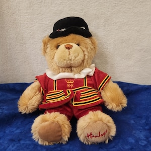 May include: A plush teddy bear wearing a red and black uniform with gold accents and a black hat. The bear has a cream-colored ruffle collar and the word "Hamley's" is embroidered on one paw.