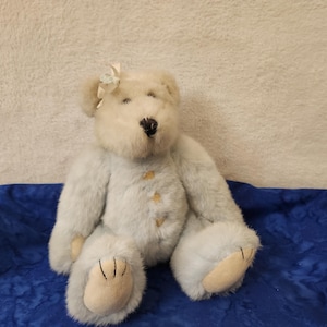 May include: A plush teddy bear with light blue fur, a cream-colored face, and a small bow on its ear. The bear has tan paws and a black nose. It is sitting on a blue textured surface.