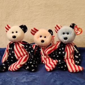 May include: Three plush teddy bears with patriotic designs. Each bear has a white face, black eyes, and a red ribbon. The bears' bodies are decorated with red and white stripes and blue stars.