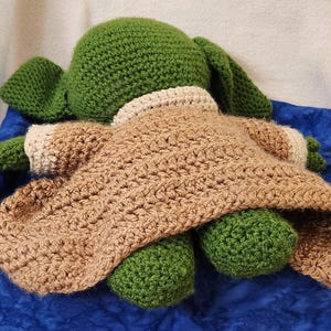 K&ouml;nnte beinhalten: Eine handgefertigte H&auml;kelpuppe, die einer beliebten Figur nachempfunden ist. Die Puppe hat einen gr&uuml;nen Kopf und Ohren, einen beigefarbenen Umhang und gr&uuml;ne F&uuml;&szlig;e. Die Puppe liegt auf einer blauen Oberfl&auml;che.