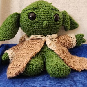 K&ouml;nnte beinhalten: Eine handgefertigte gr&uuml;ne H&auml;kelpuppe mit gro&szlig;en schwarzen Augen und &uuml;bergro&szlig;en Ohren. Die Puppe tr&auml;gt einen beigefarbenen und cremefarbenen Umhang. Die Puppe sitzt auf einer blauen Oberfl&auml;che.