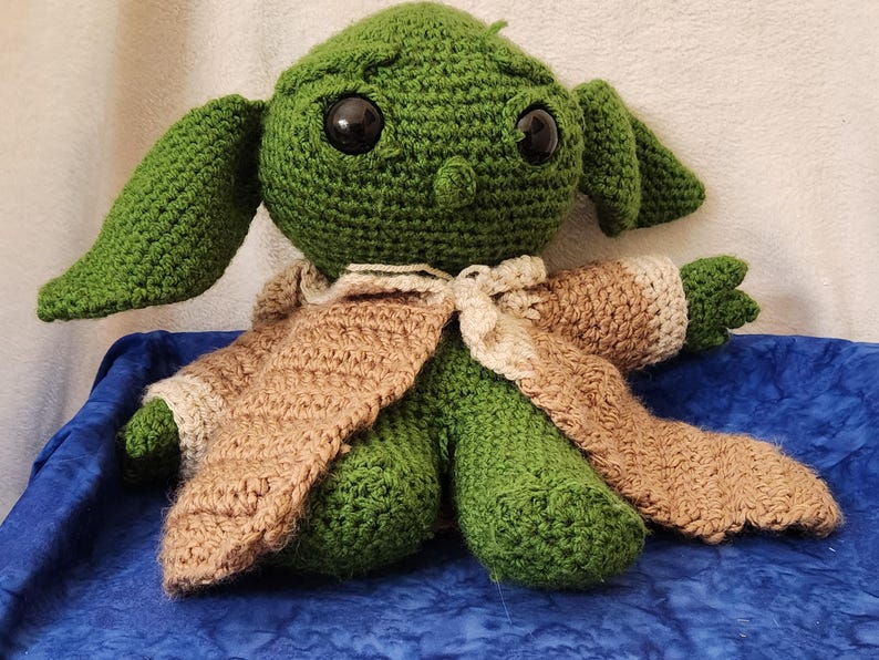 K&ouml;nnte beinhalten: Eine handgefertigte gr&uuml;ne H&auml;kelpuppe mit gro&szlig;en schwarzen Augen und &uuml;bergro&szlig;en Ohren. Die Puppe tr&auml;gt einen beigefarbenen und cremefarbenen Umhang. Die Puppe sitzt auf einer blauen Oberfl&auml;che.