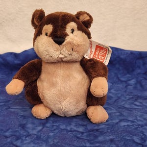 May include: A plush stuffed animal resembling a beaver with dark brown fur on the head and back, and light tan fur on the face and belly. It features a black nose, small ears, and a single visible white tooth. A white tag reads Gotta Getta GUND.