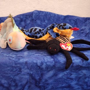May include: A collection of three plush toys including a light-colored lizard with a spiky crest, a blue and black spotted snake with a red tongue, and a black spider with orange stripes on its back, each featuring a red heart-shaped Ty tag.