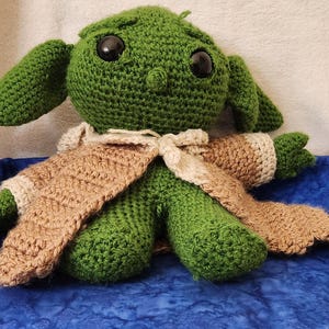 K&ouml;nnte beinhalten: Eine handgefertigte gr&uuml;ne H&auml;kelpuppe mit gro&szlig;en schwarzen Augen und langen Ohren. Die Puppe tr&auml;gt ein Outfit in Beige und Creme. Die Puppe sitzt auf einer blauen Oberfl&auml;che.