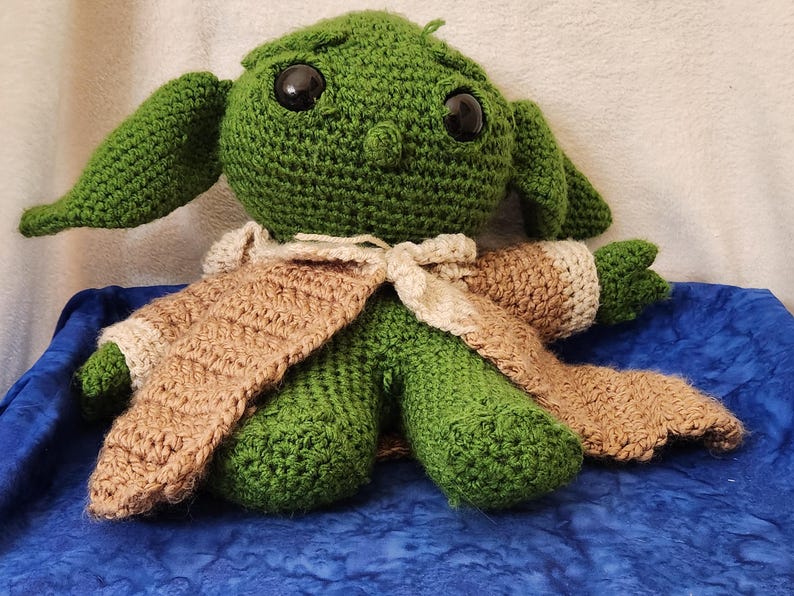 K&ouml;nnte beinhalten: Eine handgefertigte gr&uuml;ne H&auml;kelpuppe mit gro&szlig;en schwarzen Augen und langen Ohren. Die Puppe tr&auml;gt ein Outfit in Beige und Creme. Die Puppe sitzt auf einer blauen Oberfl&auml;che.