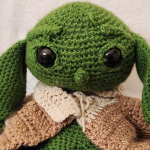 K&ouml;nnte beinhalten: Handgemachte H&auml;kelpuppe einer Figur mit gro&szlig;en schwarzen Augen, gr&uuml;ner Haut und gro&szlig;en Ohren. Die Puppe tr&auml;gt ein braunes und cremefarbenes Outfit. Die Puppe ist eine beliebte Figur aus einem Science-Fiction-Franchise.
