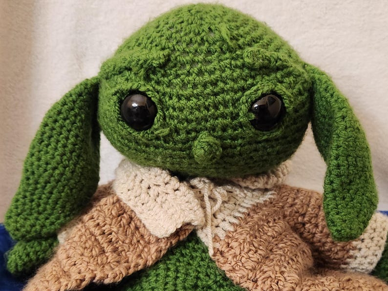 K&ouml;nnte beinhalten: Handgemachte H&auml;kelpuppe einer Figur mit gro&szlig;en schwarzen Augen, gr&uuml;ner Haut und gro&szlig;en Ohren. Die Puppe tr&auml;gt ein braunes und cremefarbenes Outfit. Die Puppe ist eine beliebte Figur aus einem Science-Fiction-Franchise.