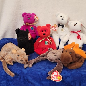 May include: A collection of nine beanie babies, including a brown rat, a black dog, a red bear, a purple bear, a white bear with a bow tie, a white bear with a red heart, and a brown moose. The beanie babies are all sitting on a blue blanket.