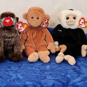 May include: Three plush toy monkeys, a brown monkey, a black monkey with a white face, and a brown and black monkey with a red face. The monkeys are sitting on a blue fabric surface.