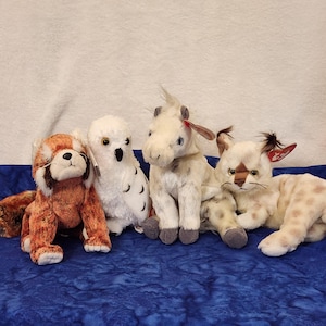 May include: Four plush animal toys are arranged on a blue surface. The toys include a red panda, a white owl, a white horse, and a spotted cat. Each toy has detailed features and is made of soft materials.