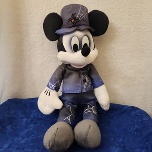 May include: A plush Mickey Mouse doll dressed in a Halloween costume. Mickey wears a dark blue jacket and pants with white accents, a matching hat with a spiderweb design, and gray shoes. The doll has black ears, a white face, and white gloves.